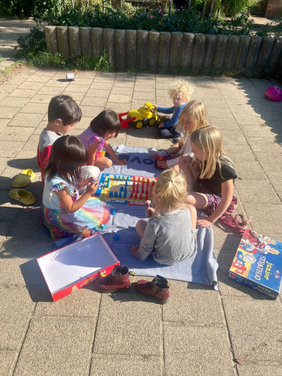 Kleurenfoto van een groepje jonge kinderen die buiten op een zonnige dag op een betegeld pleintje een bordspel spelen.