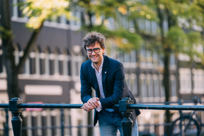 Kleurenportretfoto van Levi van Dam. Hij kijkt lachend de camera in, met zijn onderarmen steunen op een brugleuning. Op de achtergrond zijn gebouwen en bomen te zien.