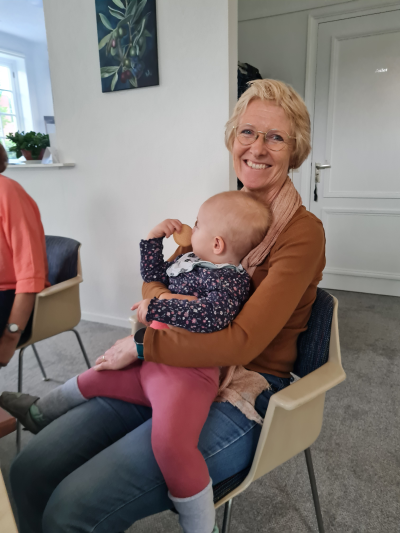 Kleurenfoto van opvoedheld Sandra. Ze zit breed lachend op een stoel en heeft een baby op schoot. met een baby op schoot.