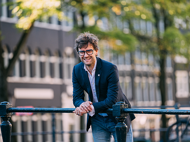 Kleurenportretfoto van Levi van Dam. Hij kijkt lachend de camera in, met zijn onderarmen steunend op een brugleuning. Op de achtergrond zijn gebouwen en bomen te zien.